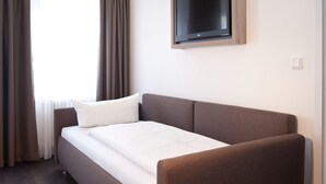 A modern hotel room with a single bed, a flat-screen TV mounted on the wall, and a window with curtains. - Manhattan Hotel (Frankfurt)