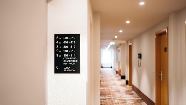 A hotel hallway with a sign listing room numbers and their corresponding amenities, including a coworking conference restroom and lobby restroom. - City Park Hotel (Frankfurt an der Oder)