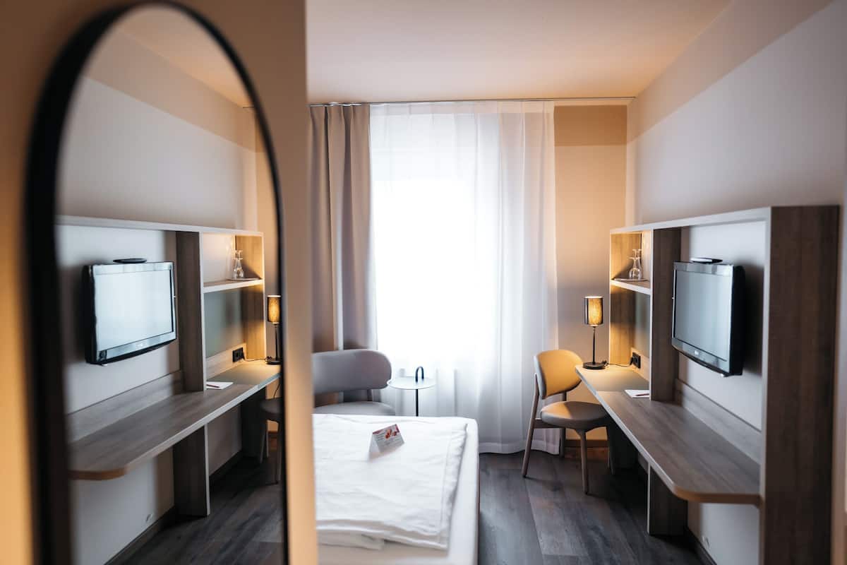 Una habitación de hotel con una cama, dos mesitas de noche, un escritorio, una silla, un televisor y un espejo.