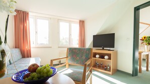 A living room with a sofa, a chair, a TV, a table with a fruit bowl, and a window with curtains. - Apart Hotel Gera (Gera)