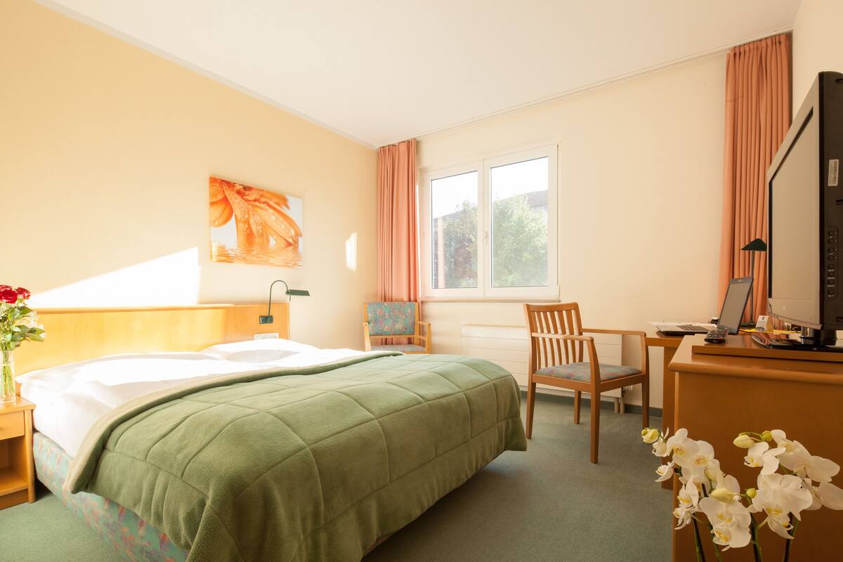 A hotel room with a bed, a desk, a chair, a window, and a painting on the wall.
