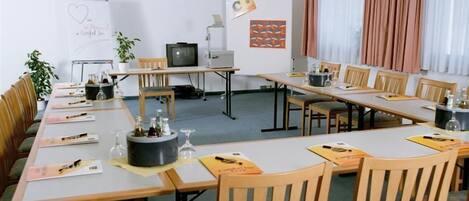 Un salón de conferencias con mesas largas, sillas, una pizarra, un televisor y un proyector.