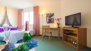 A hotel room with a bed, a desk, a television, a bottle of champagne, and a small shelf with glasses. - Apart Hotel Gera (Gera)