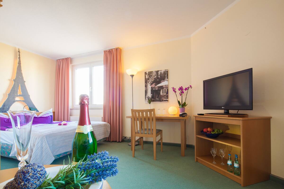A hotel room with a bed, a desk, a television, a bottle of champagne, and a small shelf with glasses.