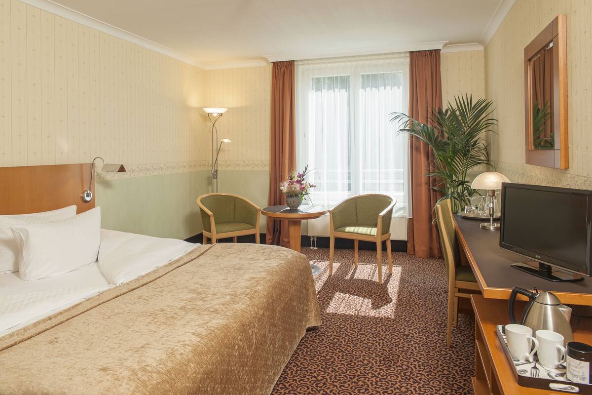 Habitación de hotel con cama, escritorio con televisor, dos sillas, una mesita con un jarrón y un ventanal con cortinas.