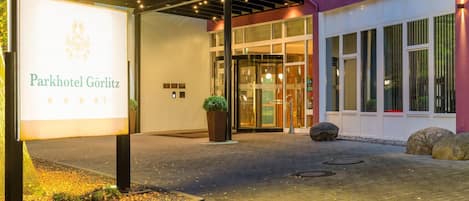 Exterior del Parkhotel Görlitz, con una entrada moderna que incluye un portal de vidrio, un área exterior cubierta con luces colgantes y un cartel.
