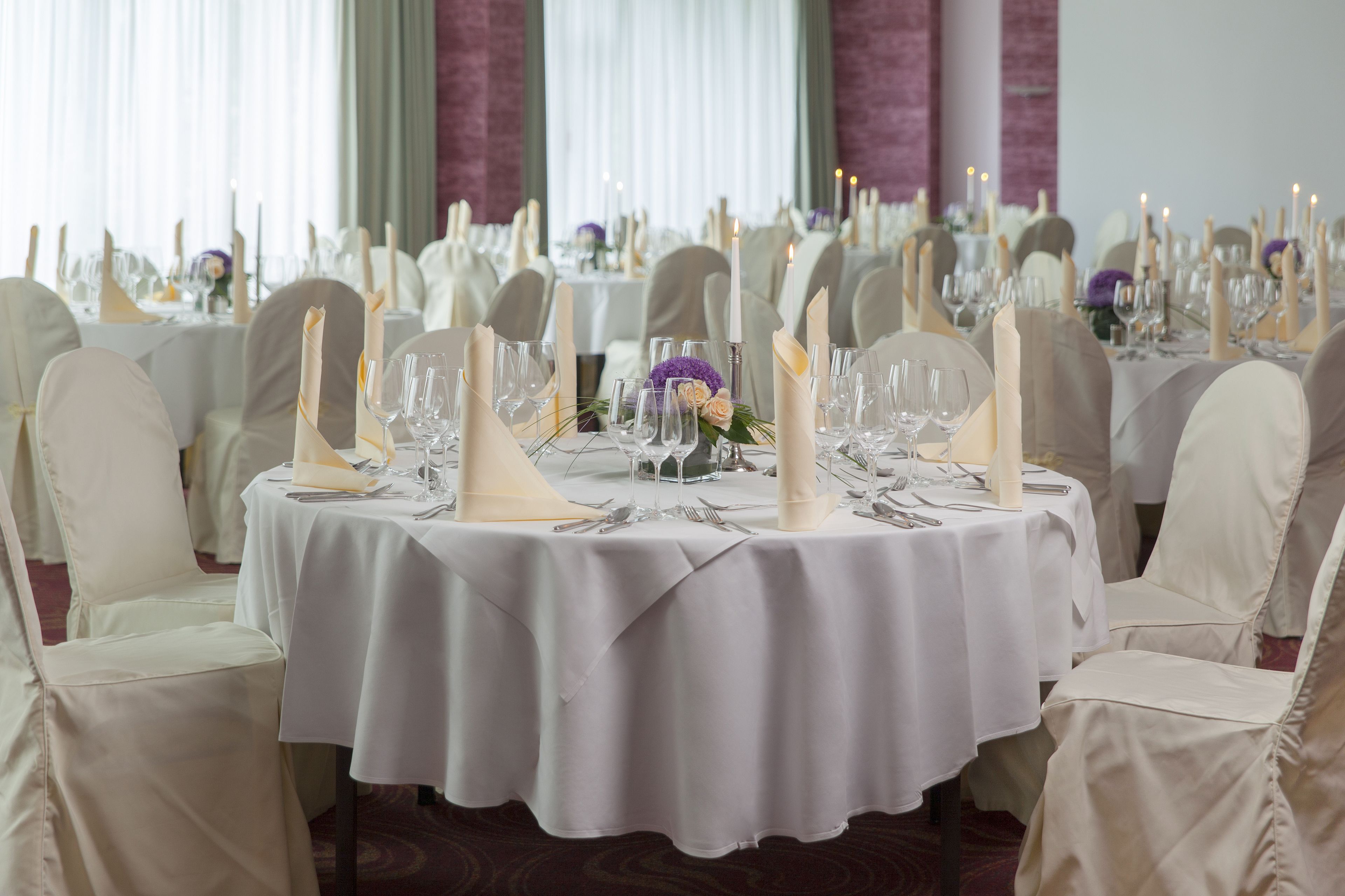 A banquet hall with round tables set for a formal event, featuring white tablecloths, candle centerpieces, and neatly arranged glassware.