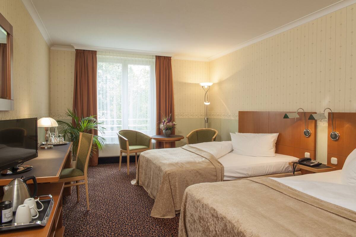 Habitación de hotel con dos camas, un escritorio con computadora, una silla, una lámpara y un ventanal con cortinas.