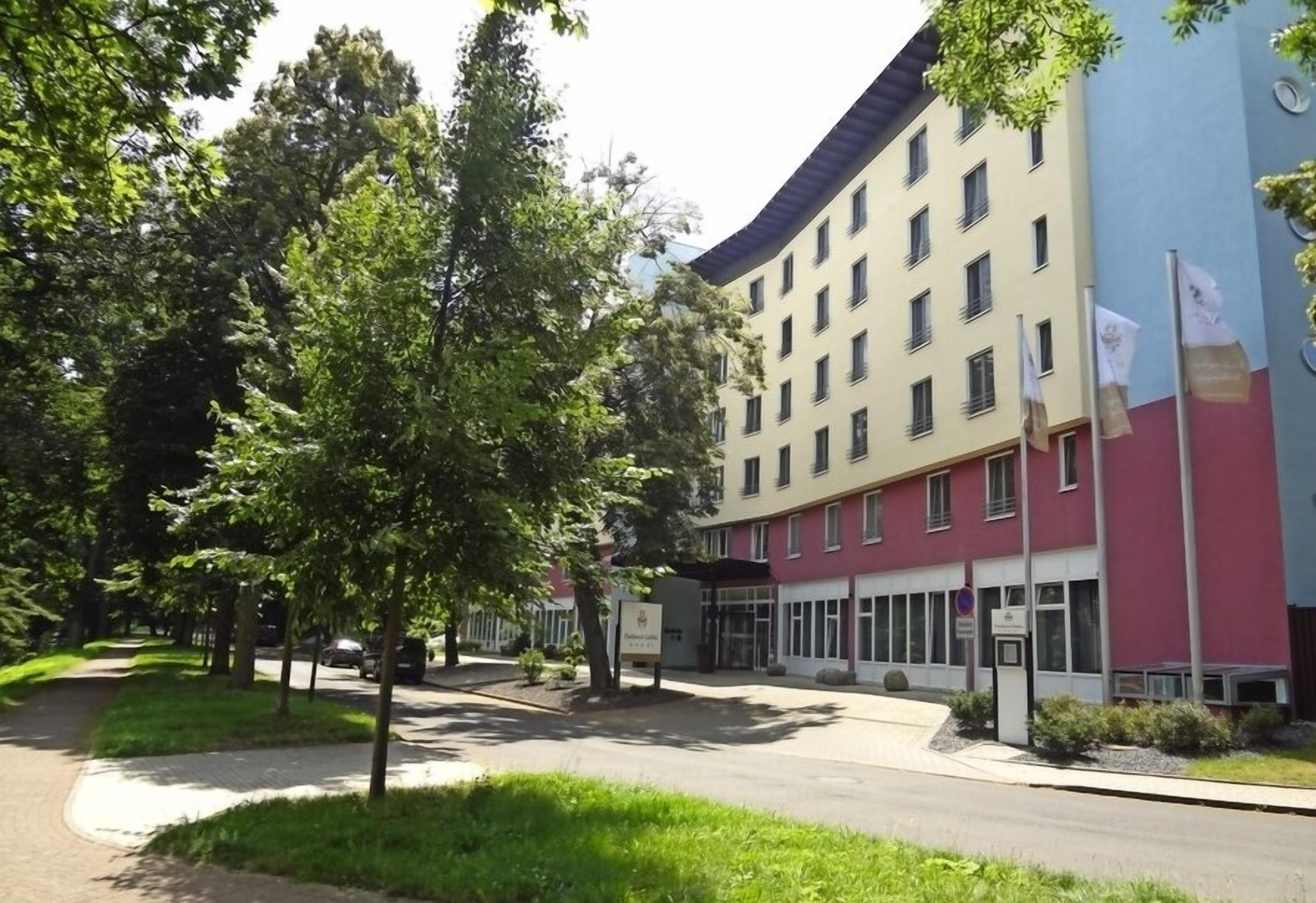 Un hotel de varios pisos con fachada roja y color crema, una entrada central y un estacionamiento con árboles.