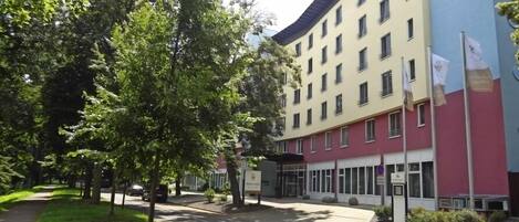 Un hotel de varios pisos con fachada roja y color crema, una entrada central y un estacionamiento con árboles.