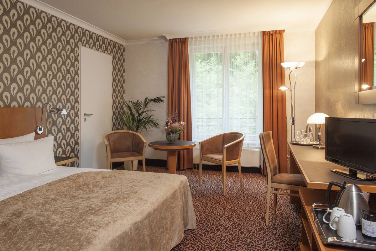 Habitación de hotel con cama, dos sillas, escritorio con televisor, cafetera y ventana con cortinas.