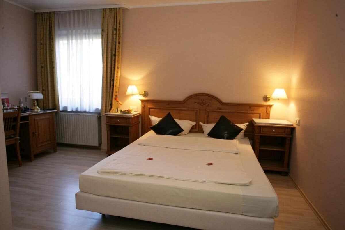 Habitación de hotel con cama doble, dos mesitas de noche con lámparas, cabecero de madera y ventana con cortinas.
