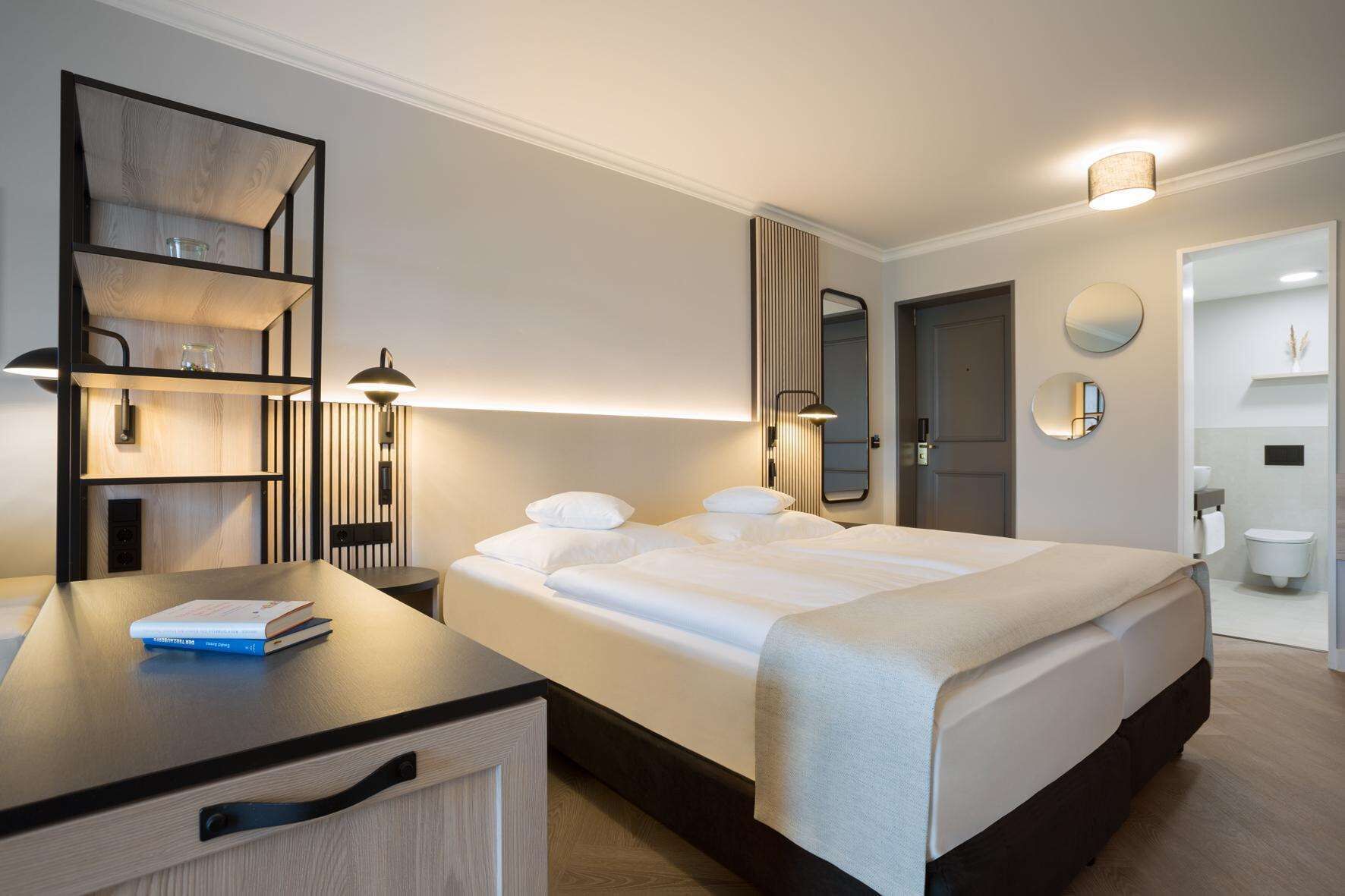 Una habitación de hotel moderna con una cama grande, un escritorio con lámpara, una estantería y un baño visible a través de una puerta abierta.