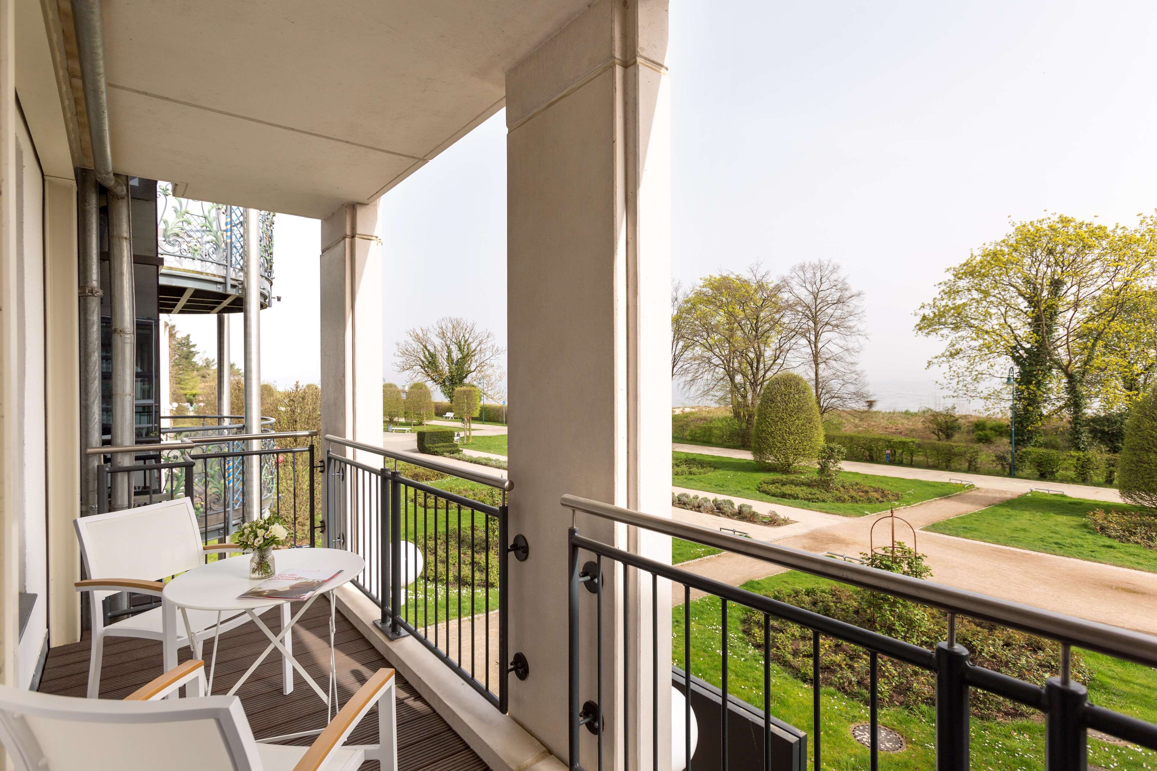 A balcony with white chairs and a table, overlooking a well-maintained garden with pathways and trees.