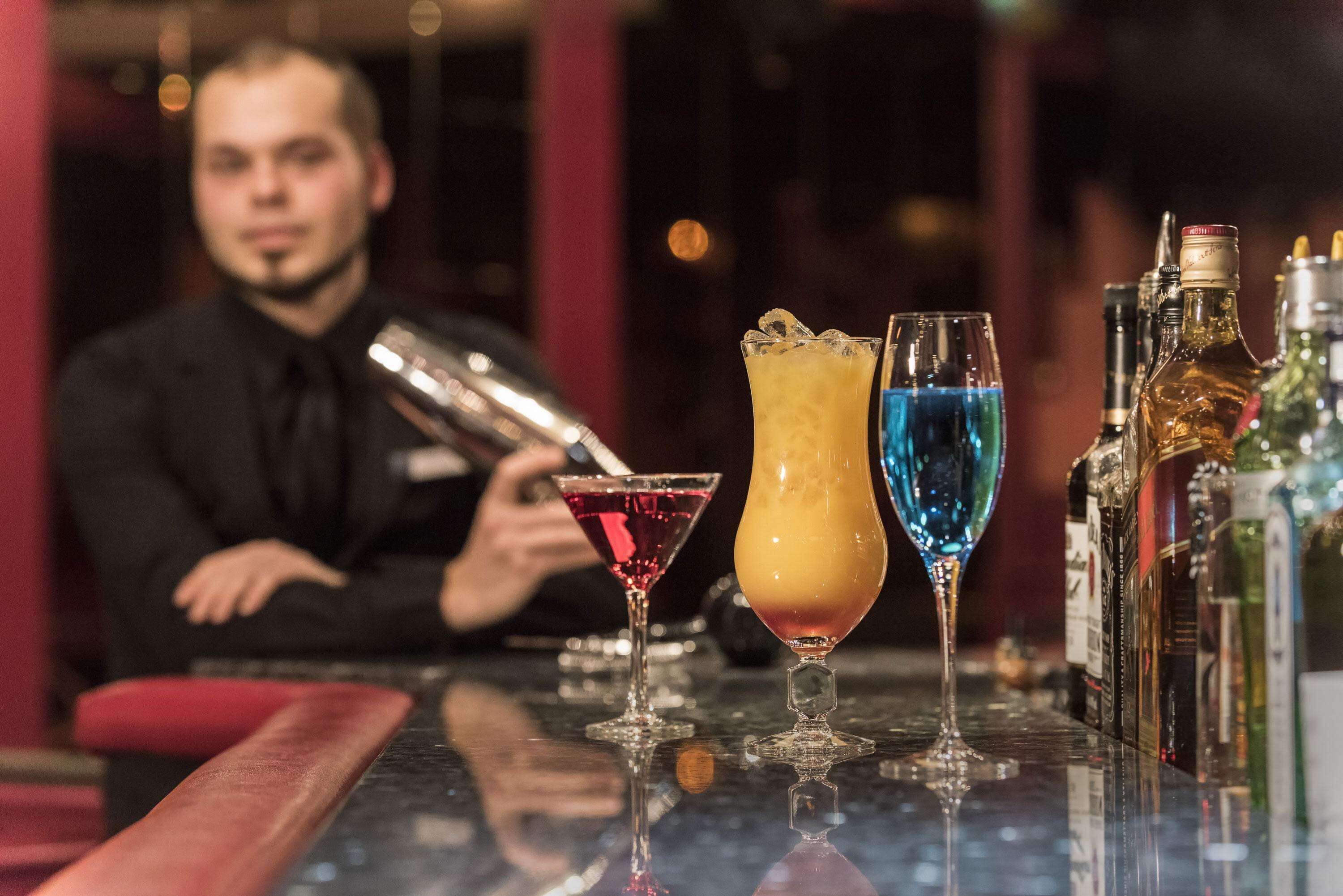 A bar with a bartender preparing drinks, a variety of cocktail glasses, and bottles of liquor on the counter.
