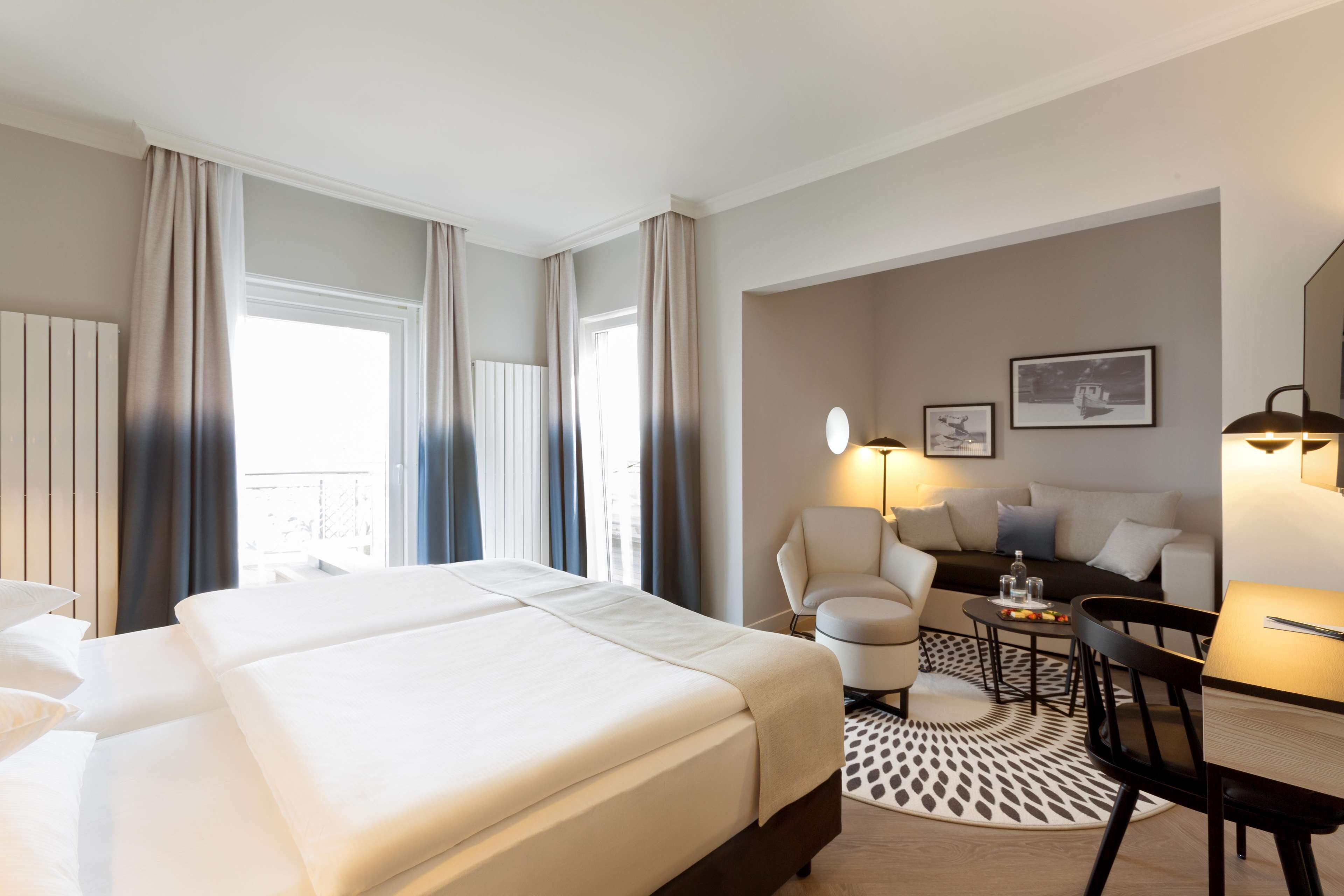 A hotel room with a large bed, a sofa, a dining table, and a chair. There is a window with curtains, a lamp, and a picture on the wall.