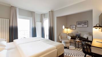 A hotel room with a large bed, a sofa, a dining table, and a chair. There is a window with curtains, a lamp, and a picture on the wall.