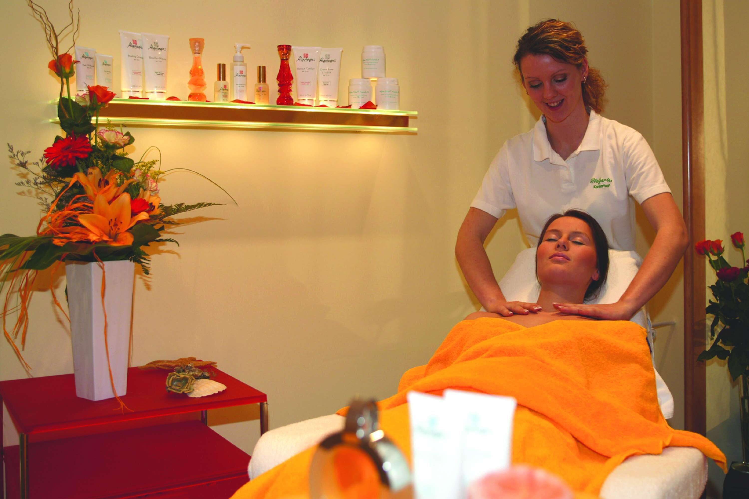Un entorno de spa con una mesa de masaje, una mesa roja con un jarrón de flores y un estante con diversos productos de spa.