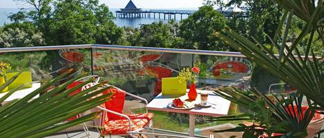 A balcony with outdoor seating, a view of a pier, and lush greenery.