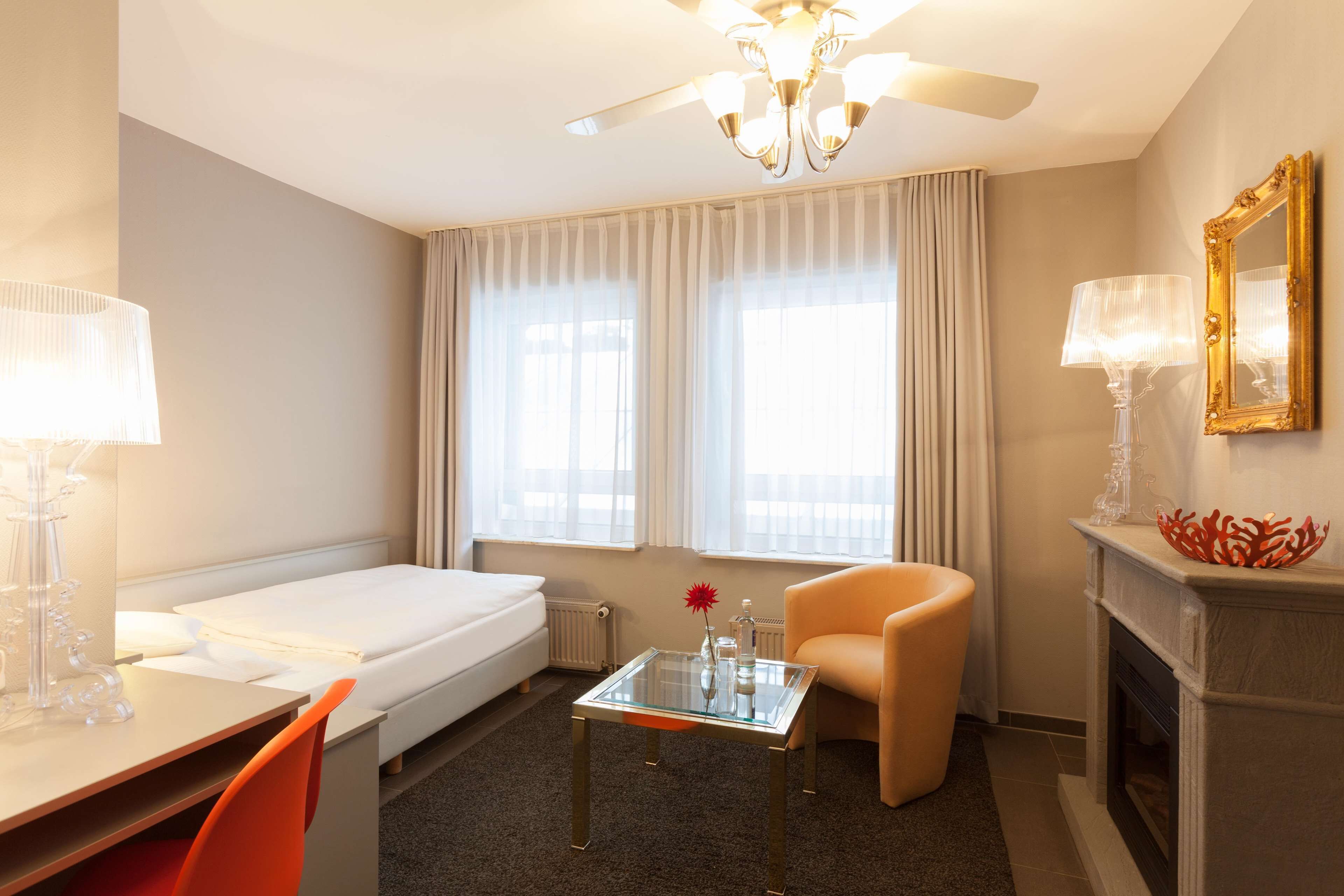 A hotel room with a bed, a glass table with a vase, a chair, a fireplace, and a large window with curtains.