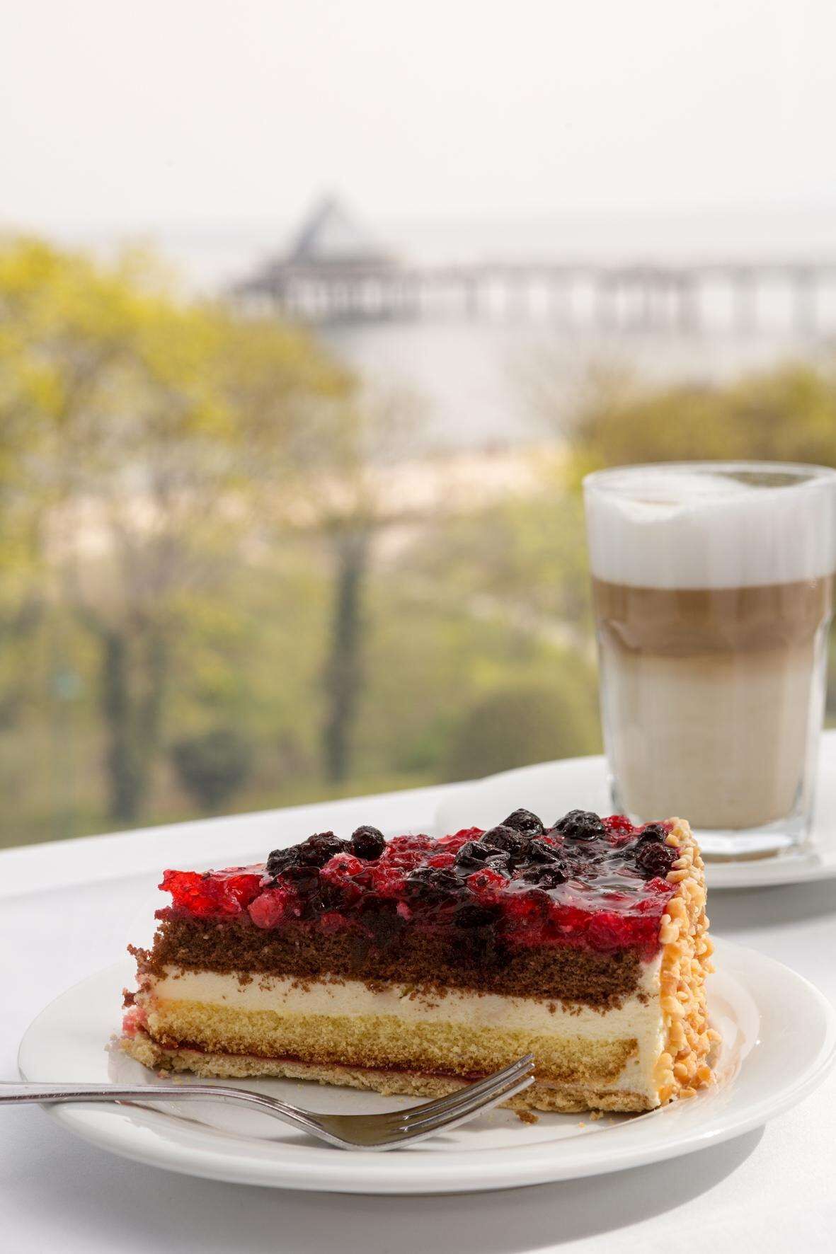 Ein Stück Schichttorte mit Beeren auf einem weißen Teller, eine Tasse Kaffee und im Hintergrund eine Brücke sowie viel Grün.