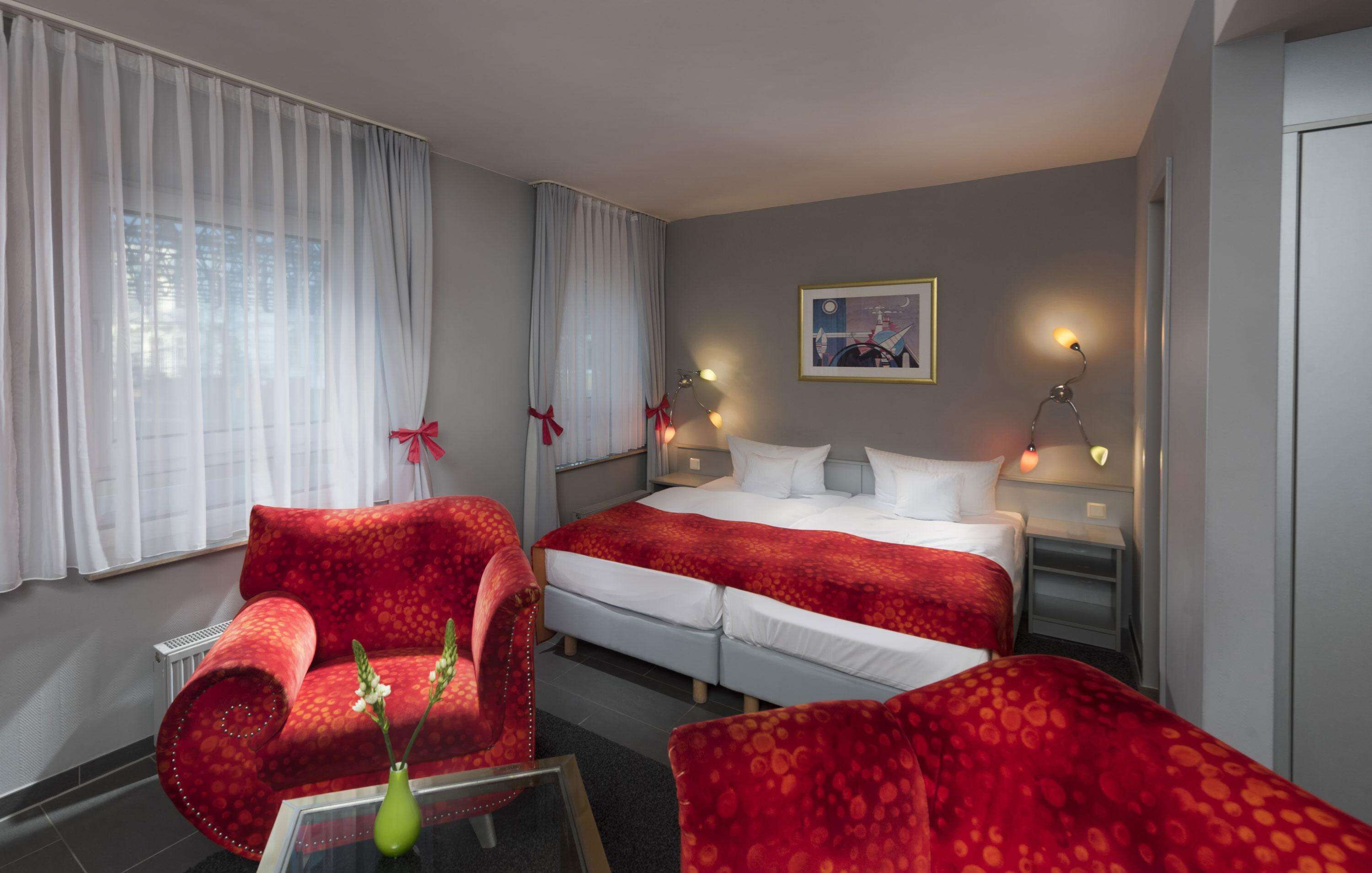 Una habitación de hotel con una cama, dos mesitas de noche, un sillón rojo, una mesa de centro de cristal con un jarrón y un cuadro en la pared.