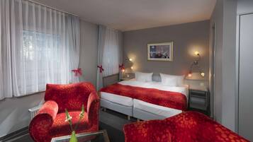 Una habitación de hotel con una cama, dos mesitas de noche, un sillón rojo, una mesa de centro de cristal con un jarrón y un cuadro en la pared.