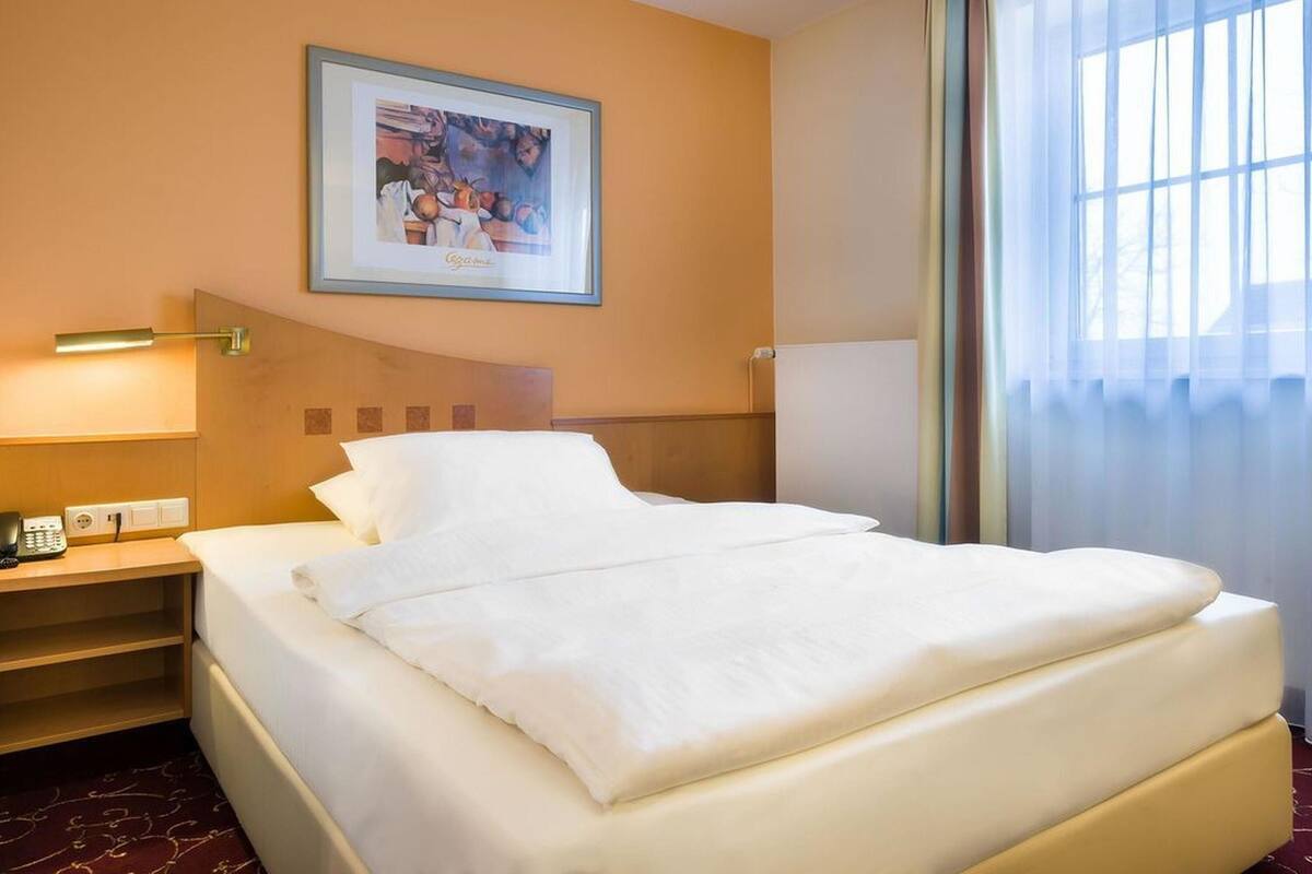 A hotel room with a bed, a bedside table, a lamp, a framed picture on the wall, and a window with curtains.