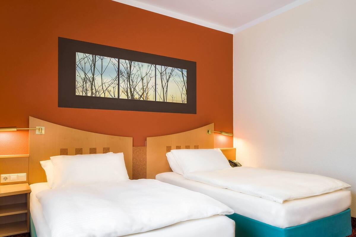 A hotel room with two beds, each with white bedding and a teal base. There are two bedside tables with lamps, and a framed picture on the wall above the beds.