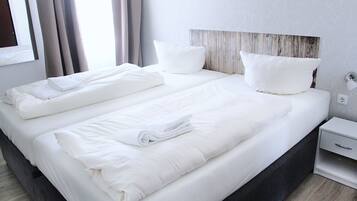 A double bed with white linens and pillows, a wooden headboard, a bedside table with a lamp, and a window with curtains.