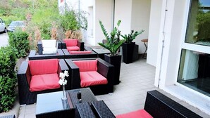 An outdoor patio with wicker furniture and red cushions, surrounded by potted plants and a view of the street. - REGIOHOTEL Leipzig West (Schkeuditz)