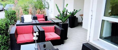 An outdoor patio with wicker furniture and red cushions, surrounded by potted plants and a view of the street.