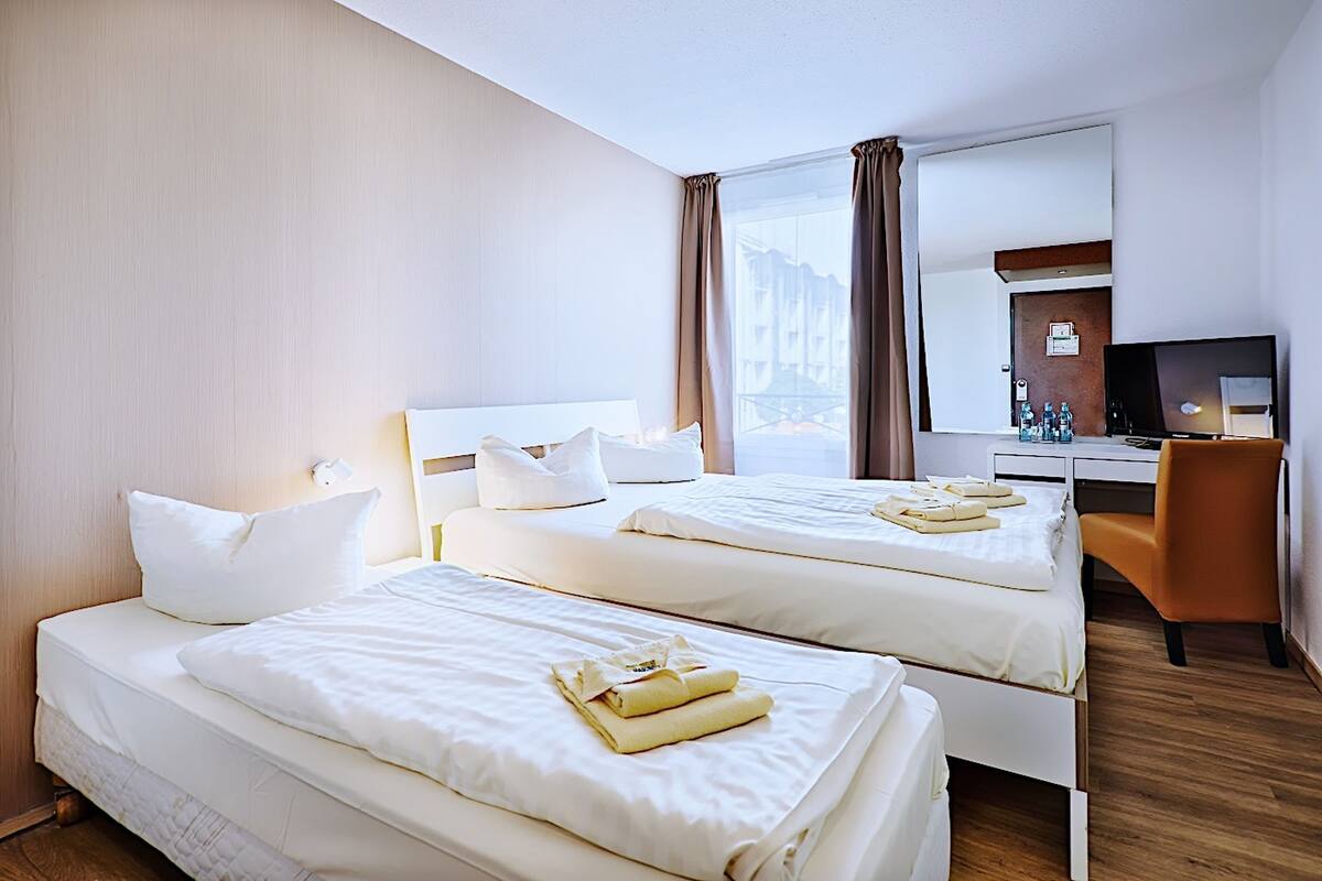A hotel room with two beds, a desk, a chair, and a large window with curtains.