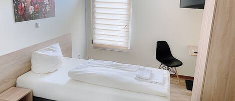 Un dormitorio ordenado con cama, mesita de noche, silla, televisor y ventana con persianas.