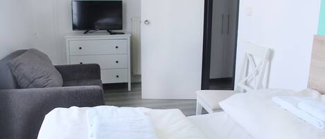 Habitación de hotel con cama, sofá, televisor sobre un tocador blanco y una puerta que da a otra habitación.