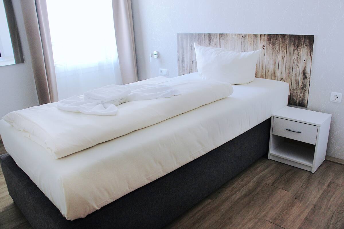 A neatly made bed with white linens and a wooden headboard, a bedside table with a drawer, and a wooden floor.