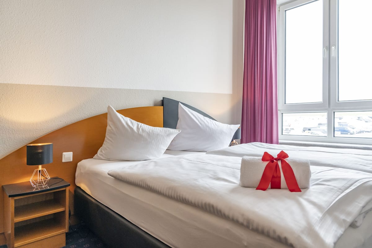 A neatly made bed with white linens and pillows, a bedside table with a lamp, and a window with curtains.