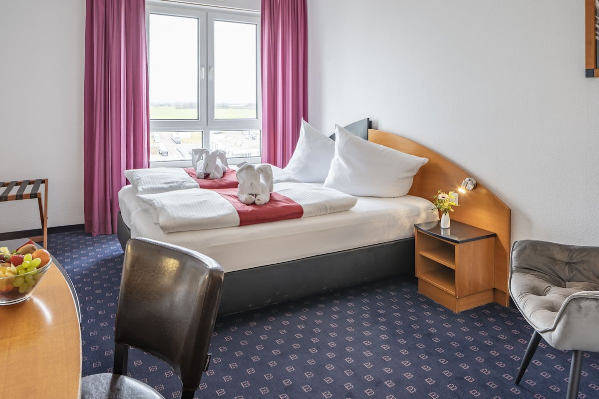 A hotel room with a bed, a bedside table with a lamp, a chair, a desk with a fruit bowl, and a window with curtains.