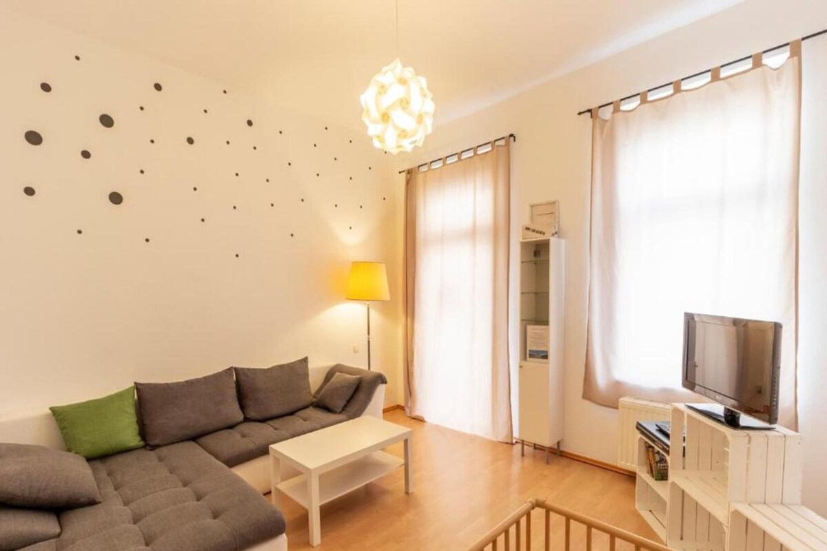 Una sala moderna con un sofá seccional, una mesita blanca, una lámpara de pie, un televisor sobre un mueble de madera y una escalera que sube a un nivel superior.
