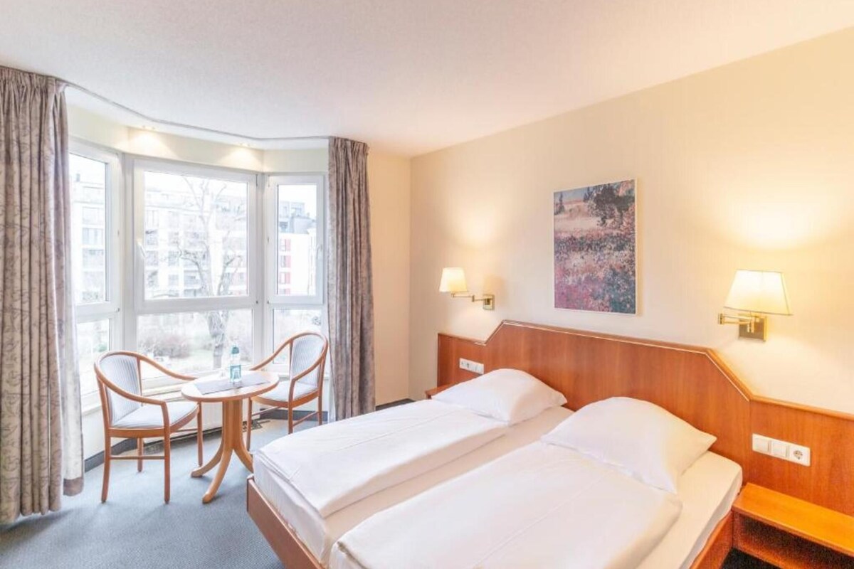 Habitación de hotel con cama, escritorio, dos sillas, ventana y un cuadro en la pared.