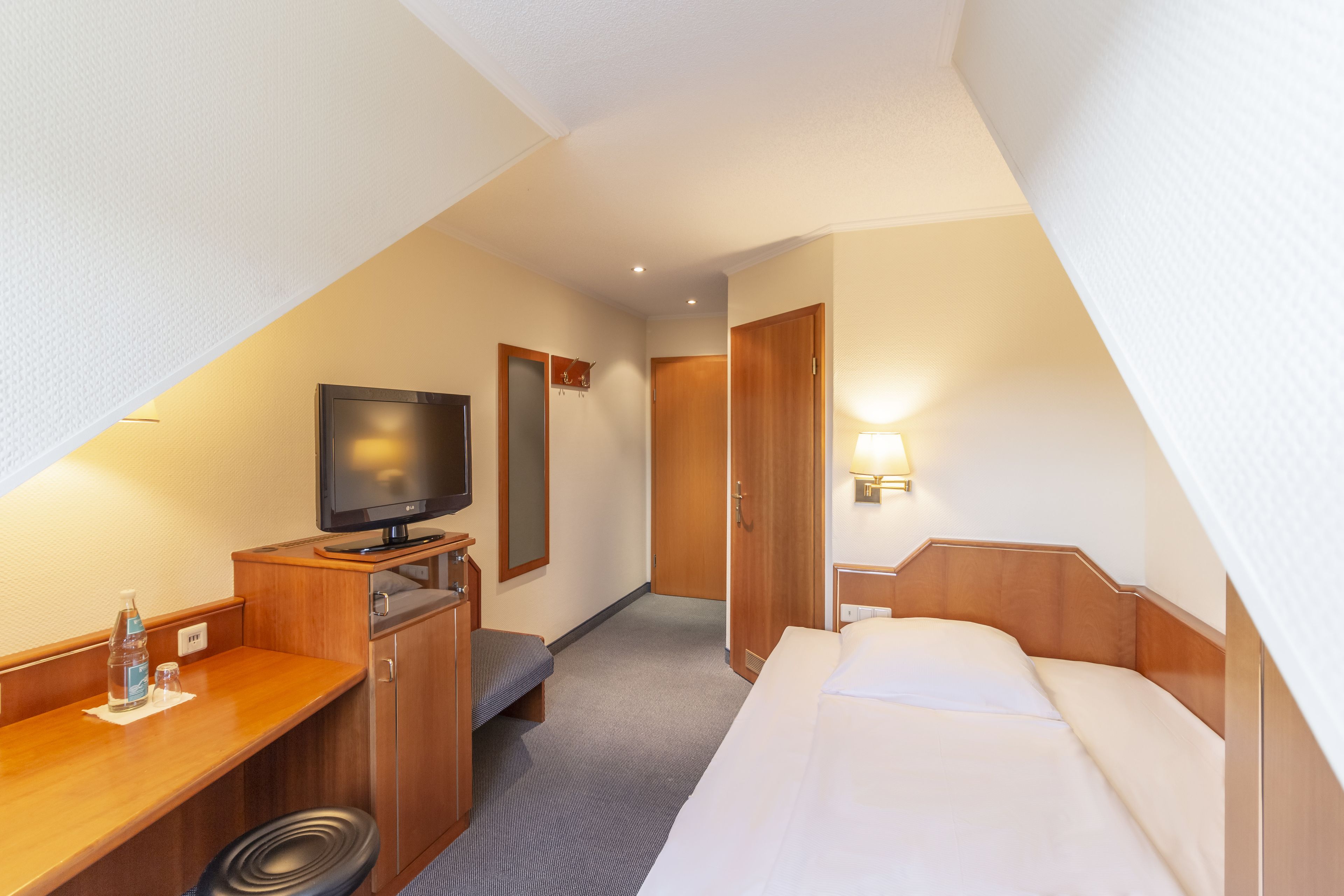 A hotel room with a bed, a desk with a television, a mirror, and a door.