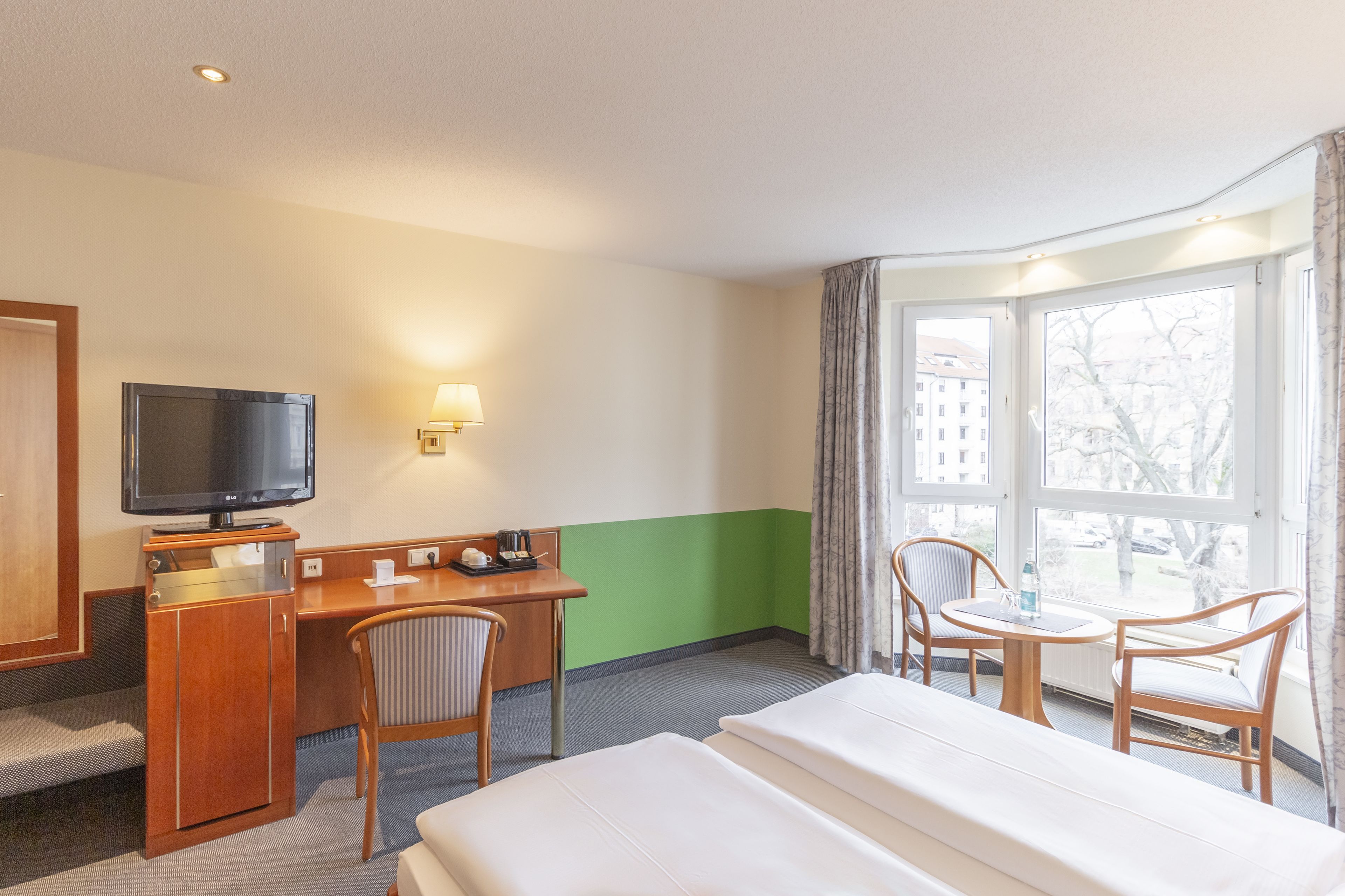 Une chambre d’hôtel comprenant un lit, un bureau avec une télévision, deux chaises, une fenêtre avec des rideaux et une vue sur les bâtiments environnants.