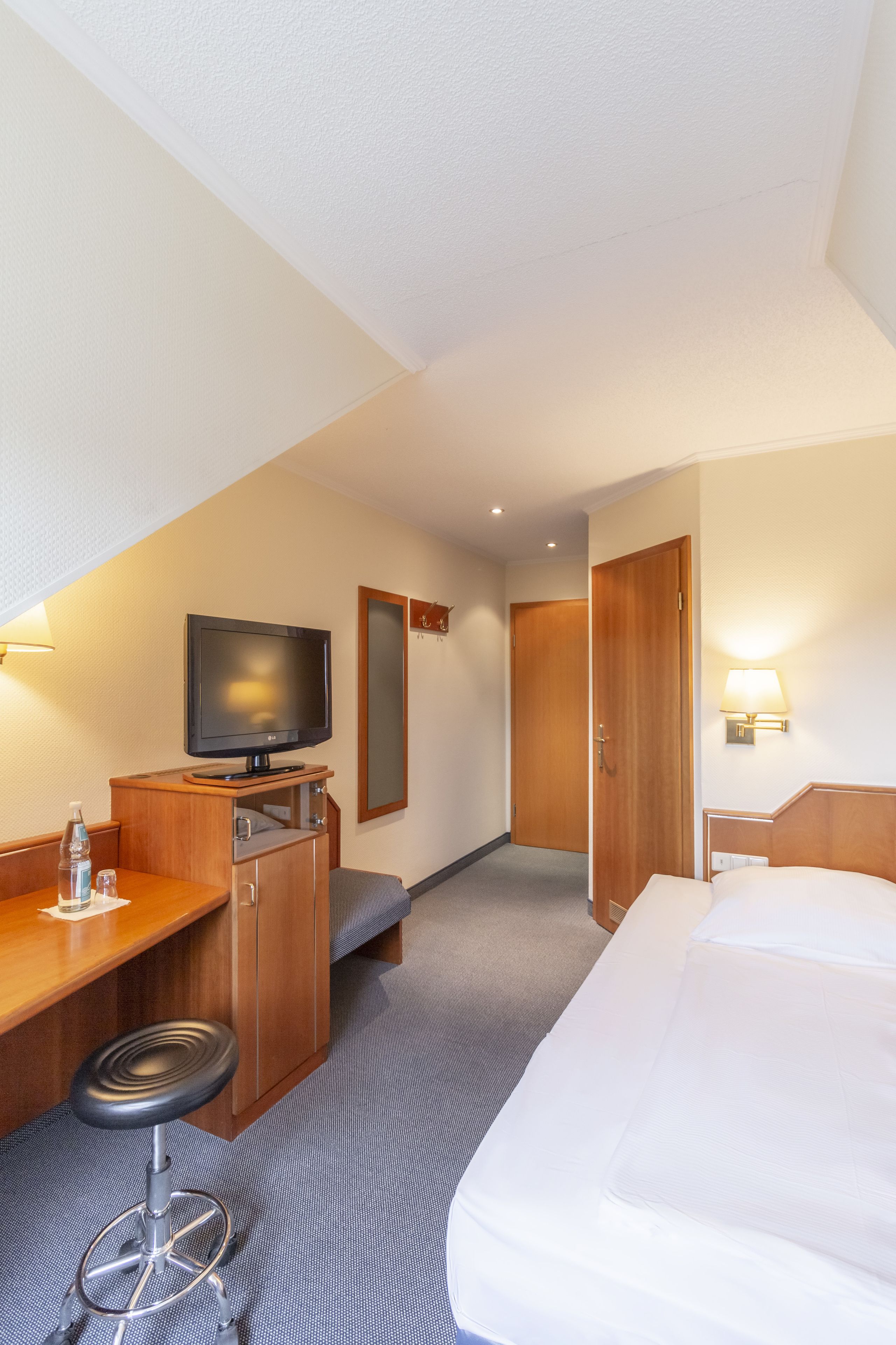 Habitación de hotel con cama, escritorio, silla, televisor, espejo y puerta.