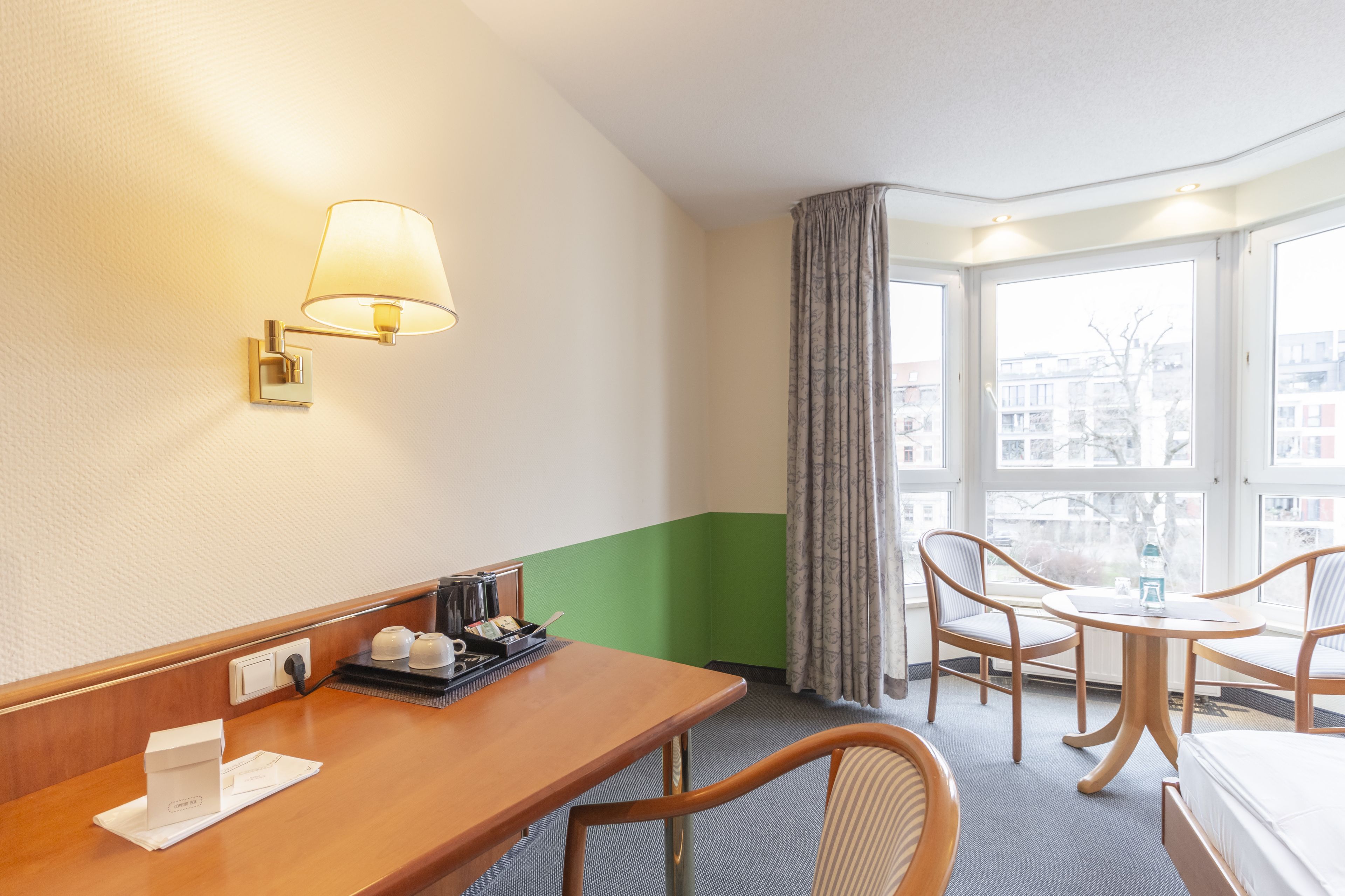 A hotel room with a wooden desk, a lamp, a bed, two chairs, a window with a view of buildings, and a green accent wall.