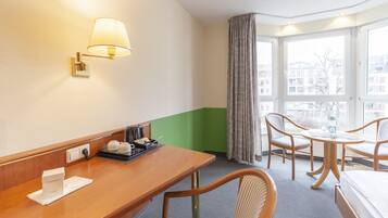 A hotel room with a wooden desk, a lamp, a bed, two chairs, a window with a view of buildings, and a green accent wall.