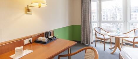 A hotel room with a wooden desk, a lamp, a bed, two chairs, a window with a view of buildings, and a green accent wall.