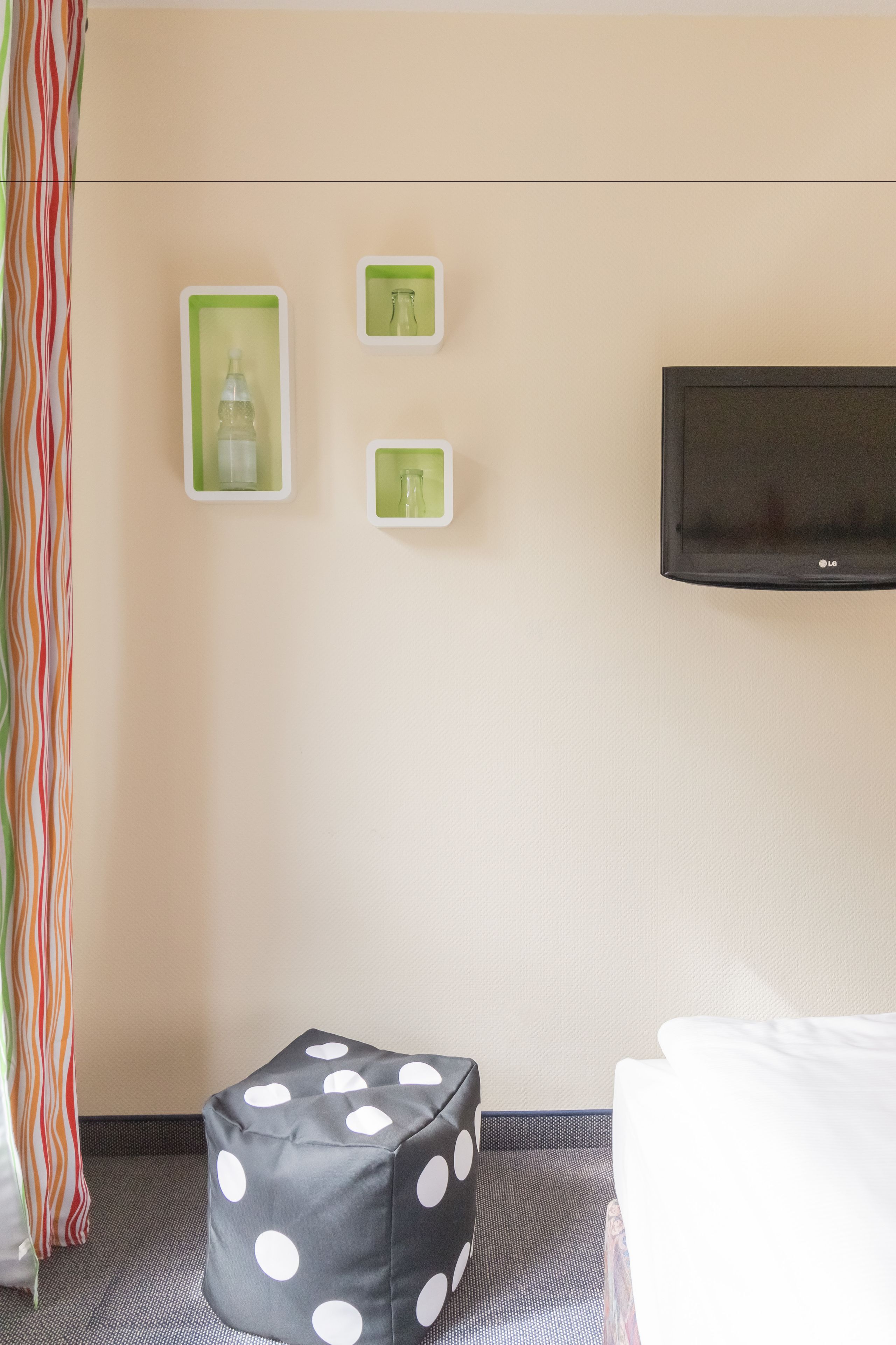A hotel room with a bed, a TV mounted on the wall, and a small seating area with a polka-dotted ottoman.