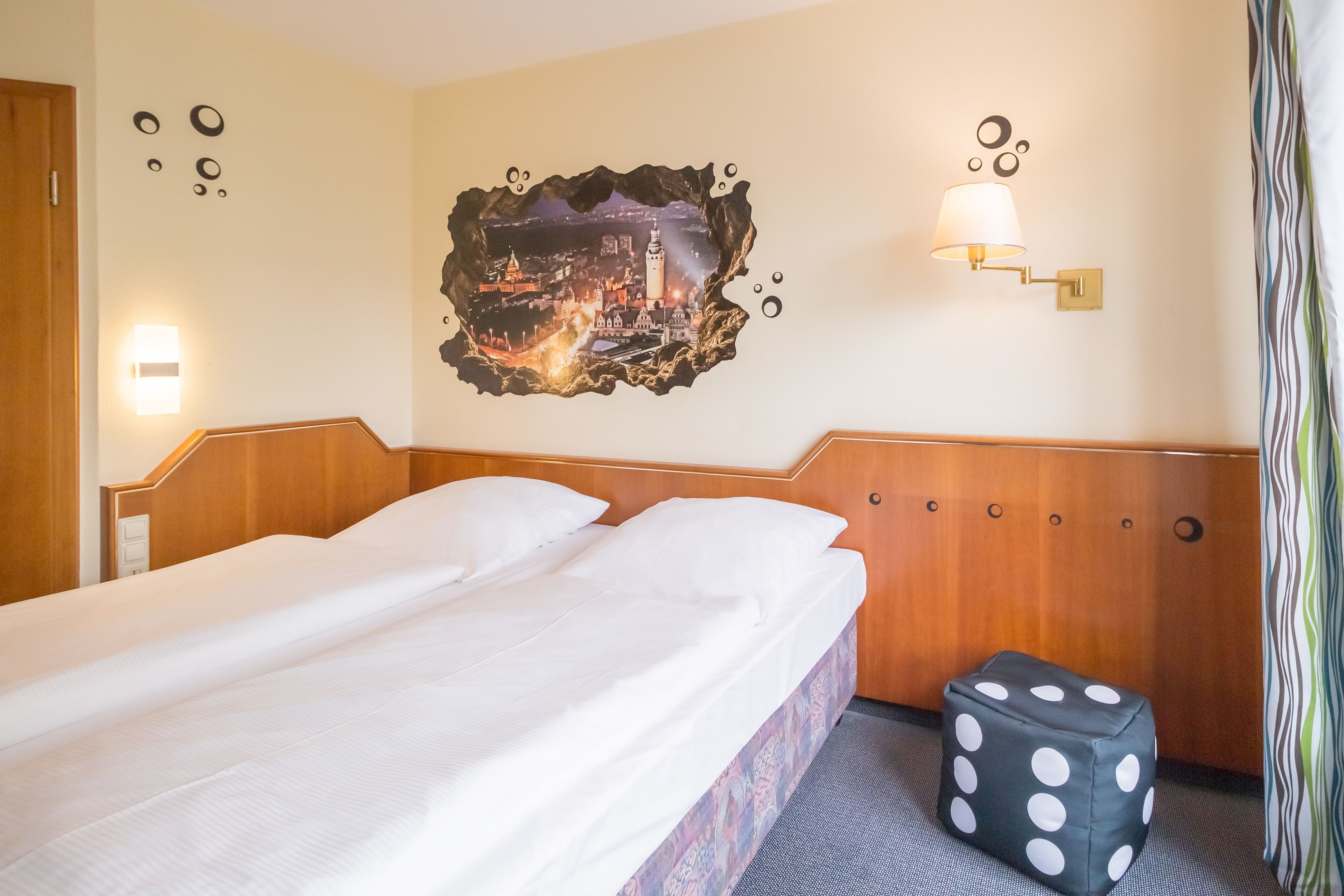 Une chambre d’hôtel avec un lit parfaitement fait, un tableau représentant un paysage urbain accroché au mur, une lampe murale et un pouf à pois.