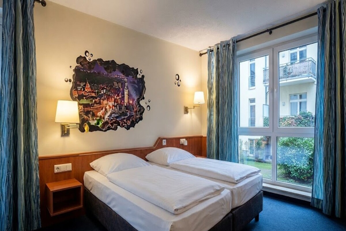 Habitación de hotel con dos camas, una mesita de noche, una lámpara de pared y un ventanal con cortinas.