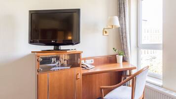 A hotel room with a wooden desk, a television, a lamp, a chair, and a window with a view.